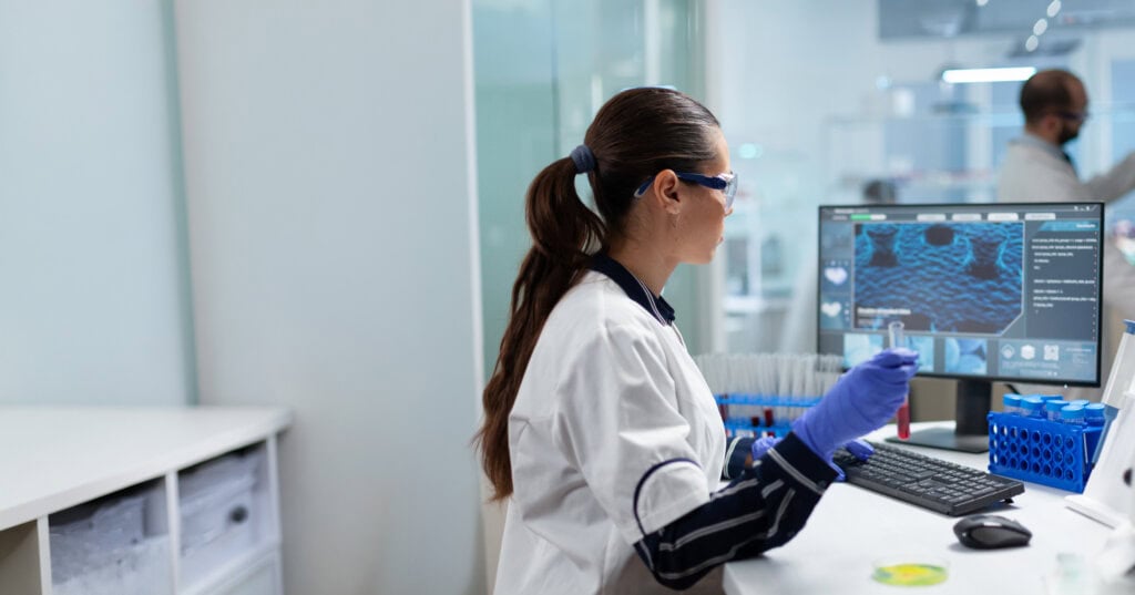 Biologin und Forscherin, die ein Blutröhrchen in der Hand hält und DNA-Analysen am Computer durchführt.